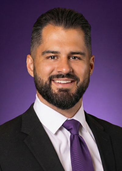 Photo of a man, smiling, wearing a white collared button up shirt, a purple tie, black jacket, with a purple background