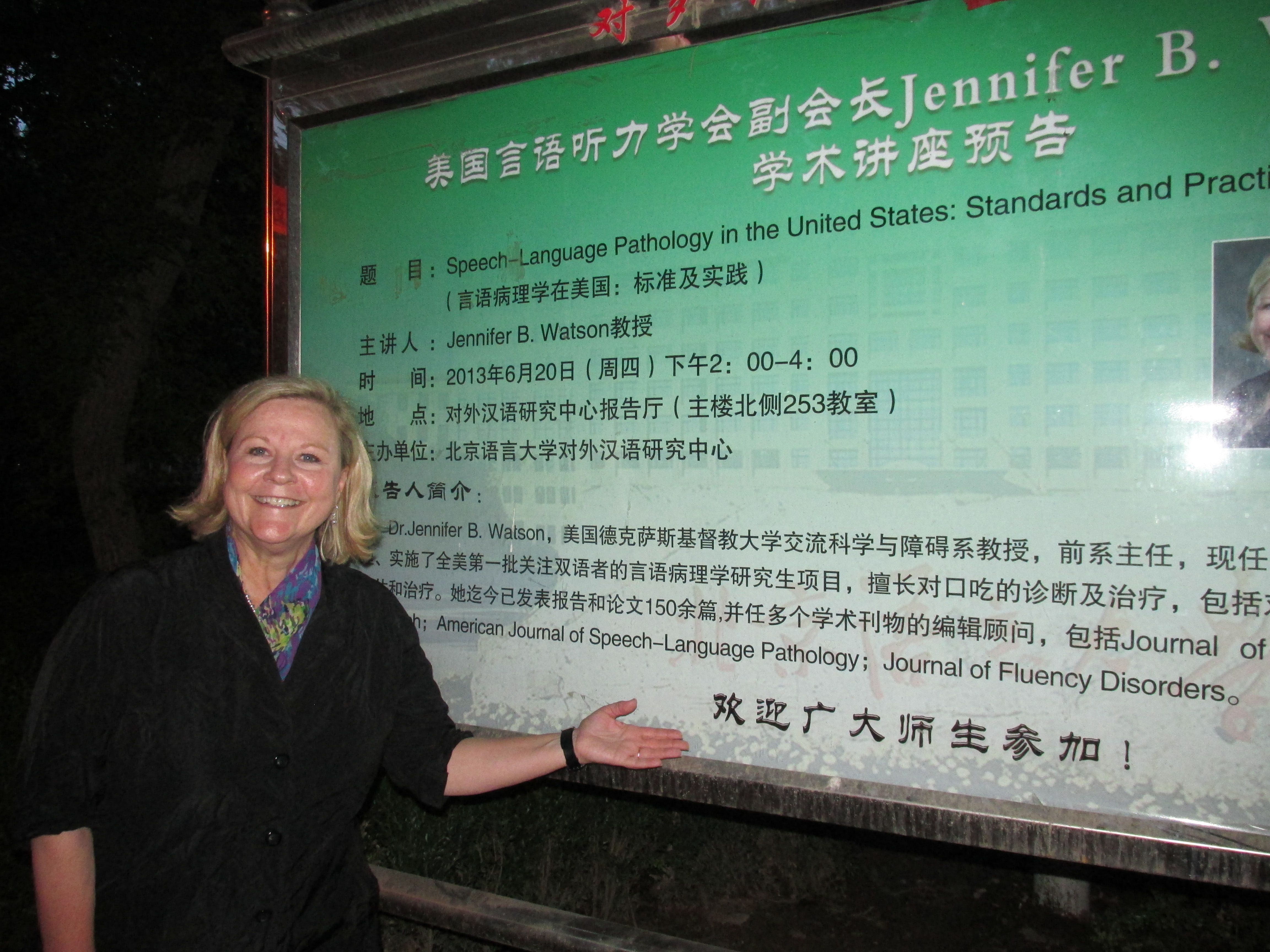 Dr. Jennifer Watson standing in front of a sign at the Beijing Language and Culture University featuring her specializing in speech-language pathology and fluency disorders.