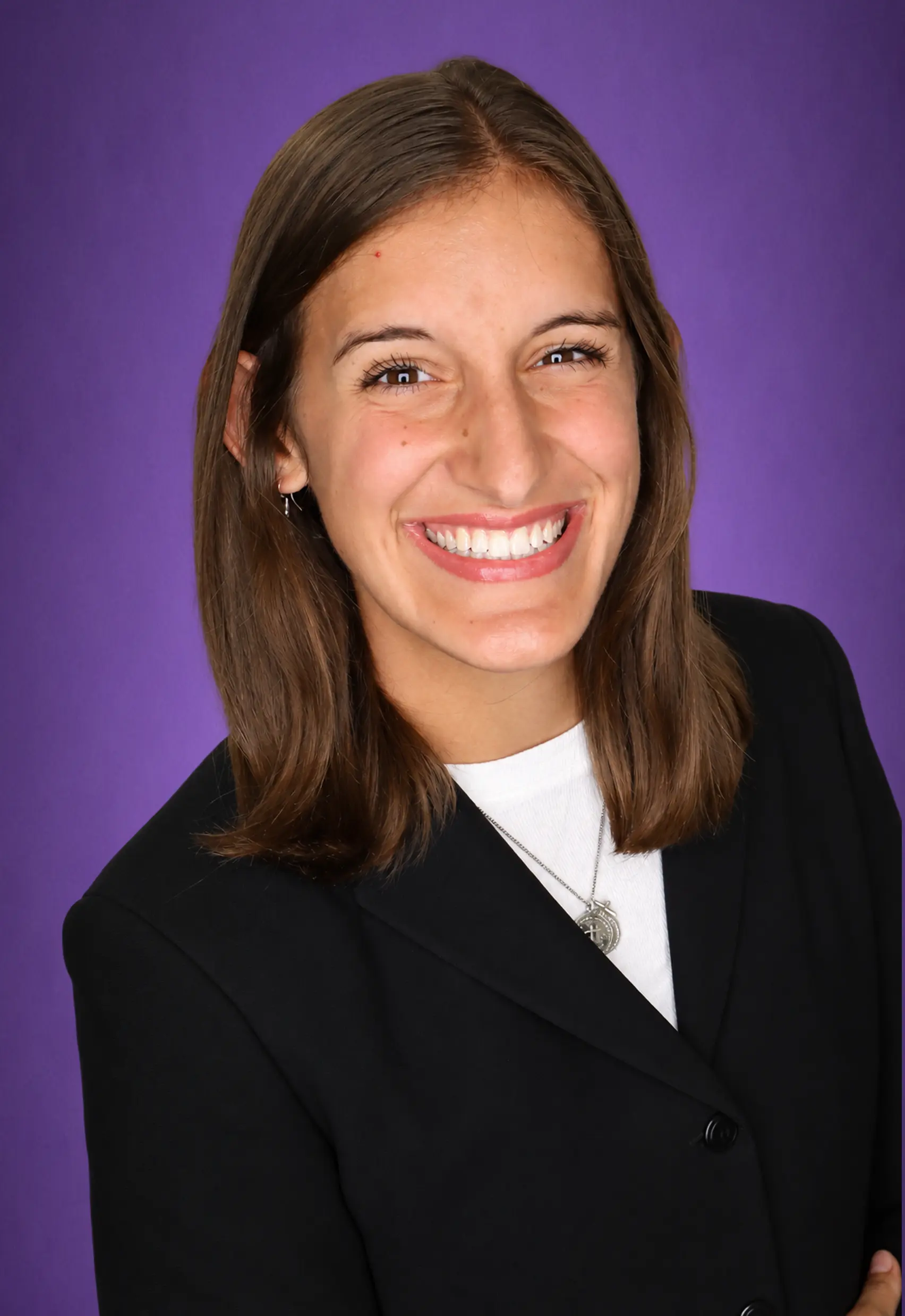 A woman, smiling, wearing a white shirt with a black blazer buttoned up, with a purple background