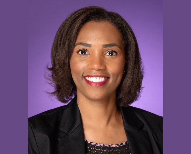 Photo of a woman smiling, wearing a multi-colored pink and purple floral shirt with a black blazer, with a purple background.