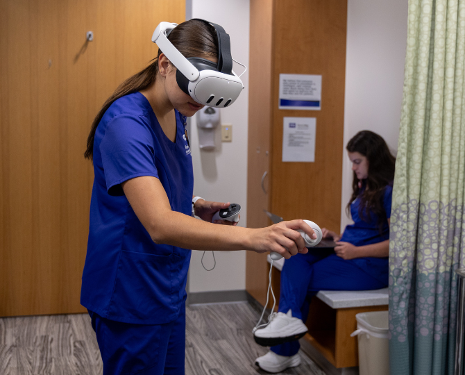 A Harris College nursing student wears a VR headset in an exam room for a virtual patient simulation exercise, with a second nursing student overseeing the simulation on a laptop.